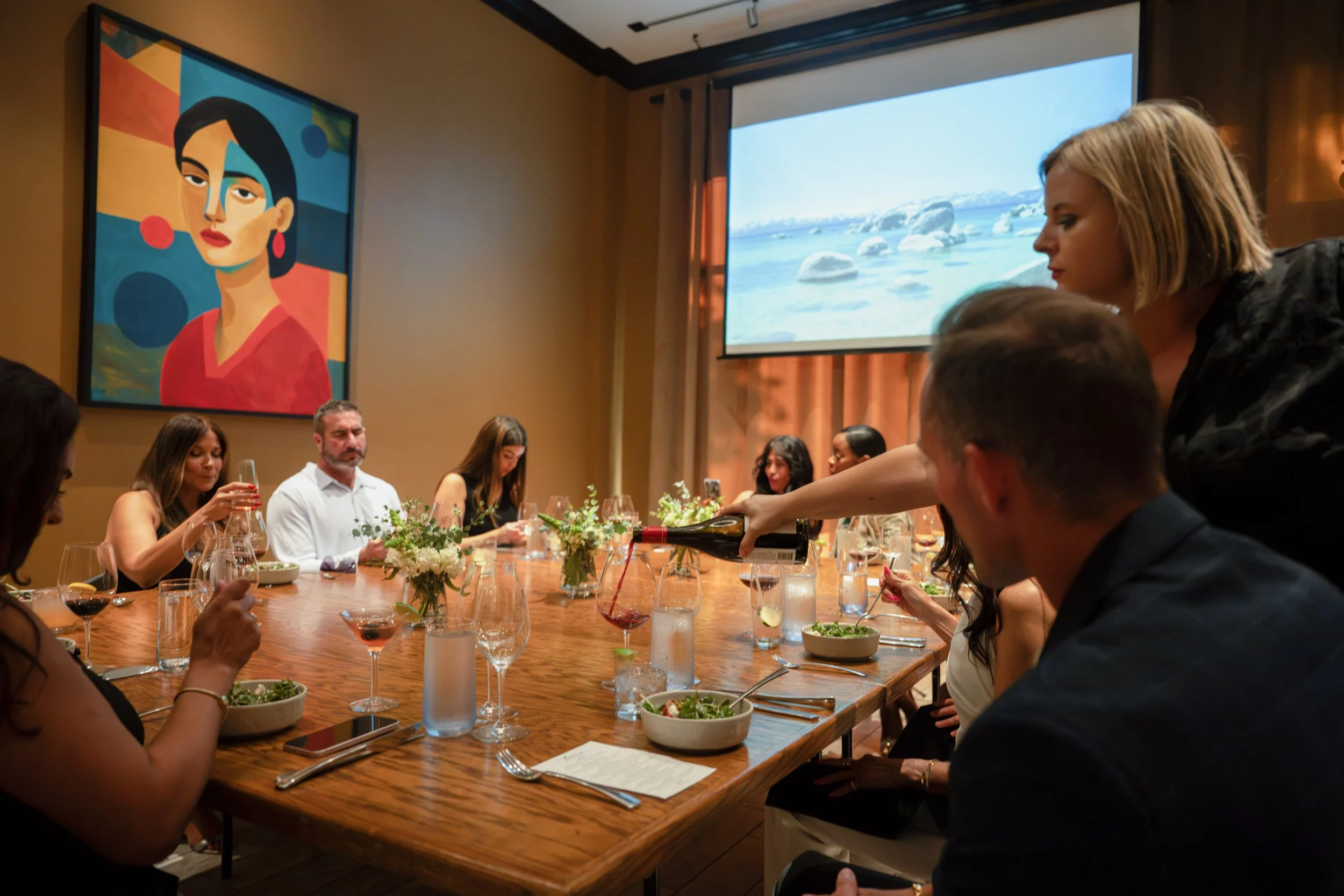 Guests seated around a long communal table during a private dinner in The Annex, with wine service and presentation screen in the background.