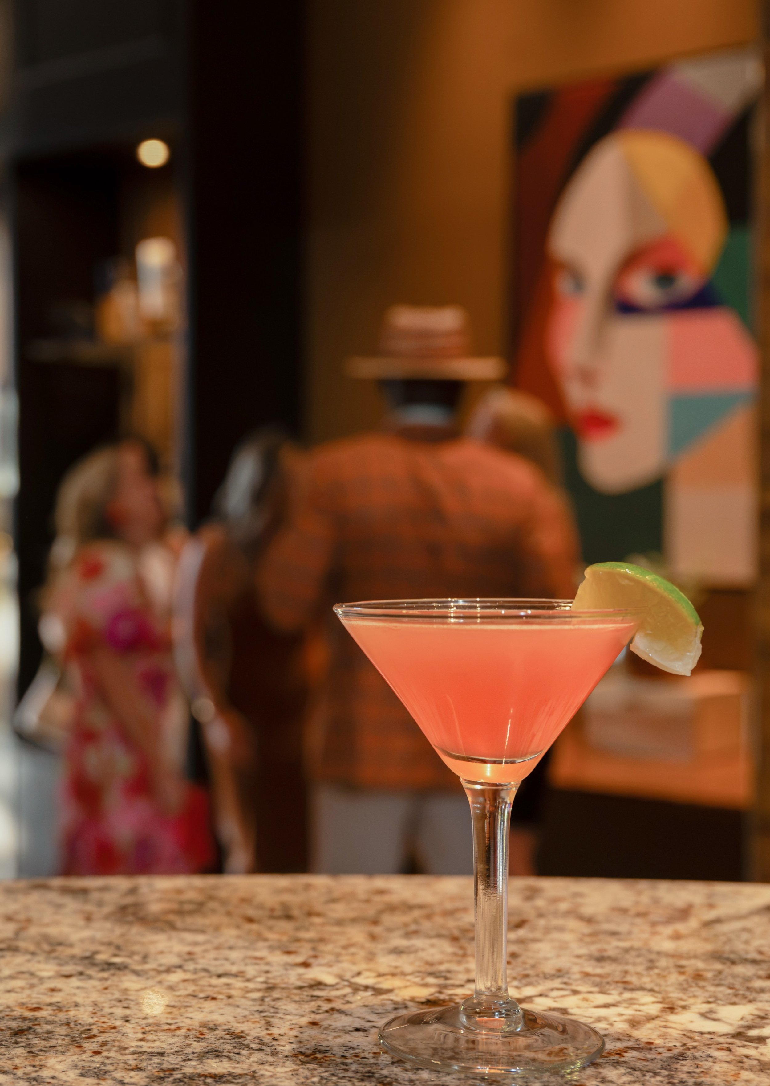 Pink cocktail in a martini glass with a lemon wedge on a granite bar counter, blurred background of people and colorful artwork.