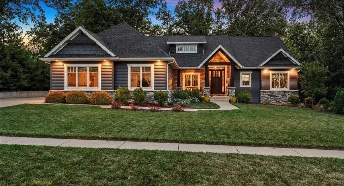 A large, dark-colored house with a gabled roof, illuminated exterior lights, and a landscaped front yard with neatly trimmed grass and shrubs, at dusk.