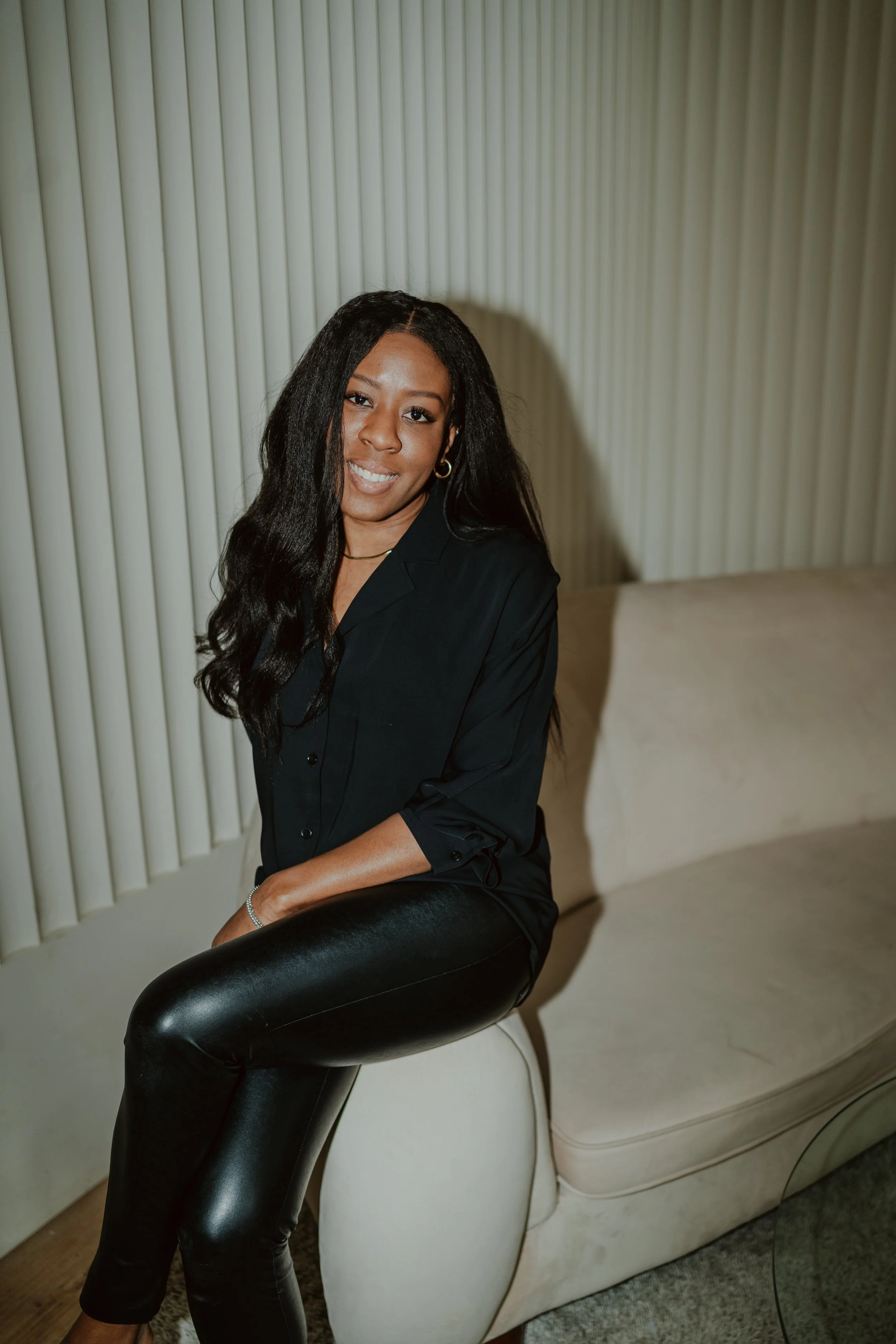 A woman with long dark hair, gold hoop earrings, and a black shirt, seated on a cream-colored chair with a draped back, in front of vertically decorated wall. She is an interior designer freelancing her services.