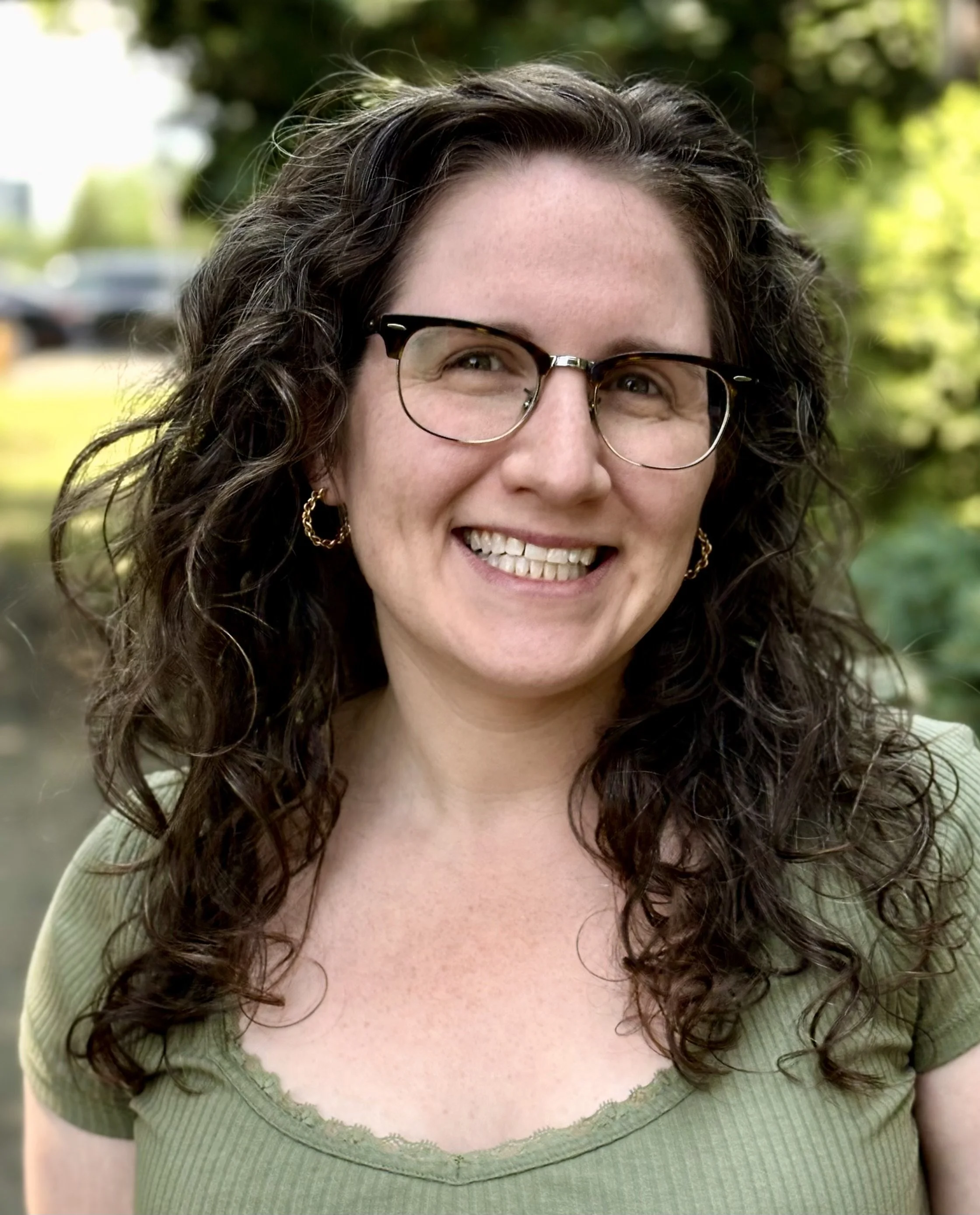 Close-up of a woman with curly brown hair, wearing glasses and smiling, outdoors with blurred green trees in the background.