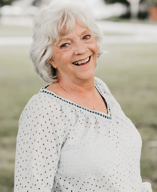 A smiling elderly woman with white hair, wearing a white eyelet blouse, standing outdoors with a grassy field in the background.