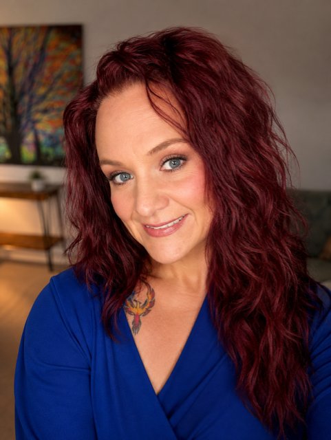 A woman with curly red hair and blue eyes, wearing a blue top, smiling indoors with a modern living room in the background.