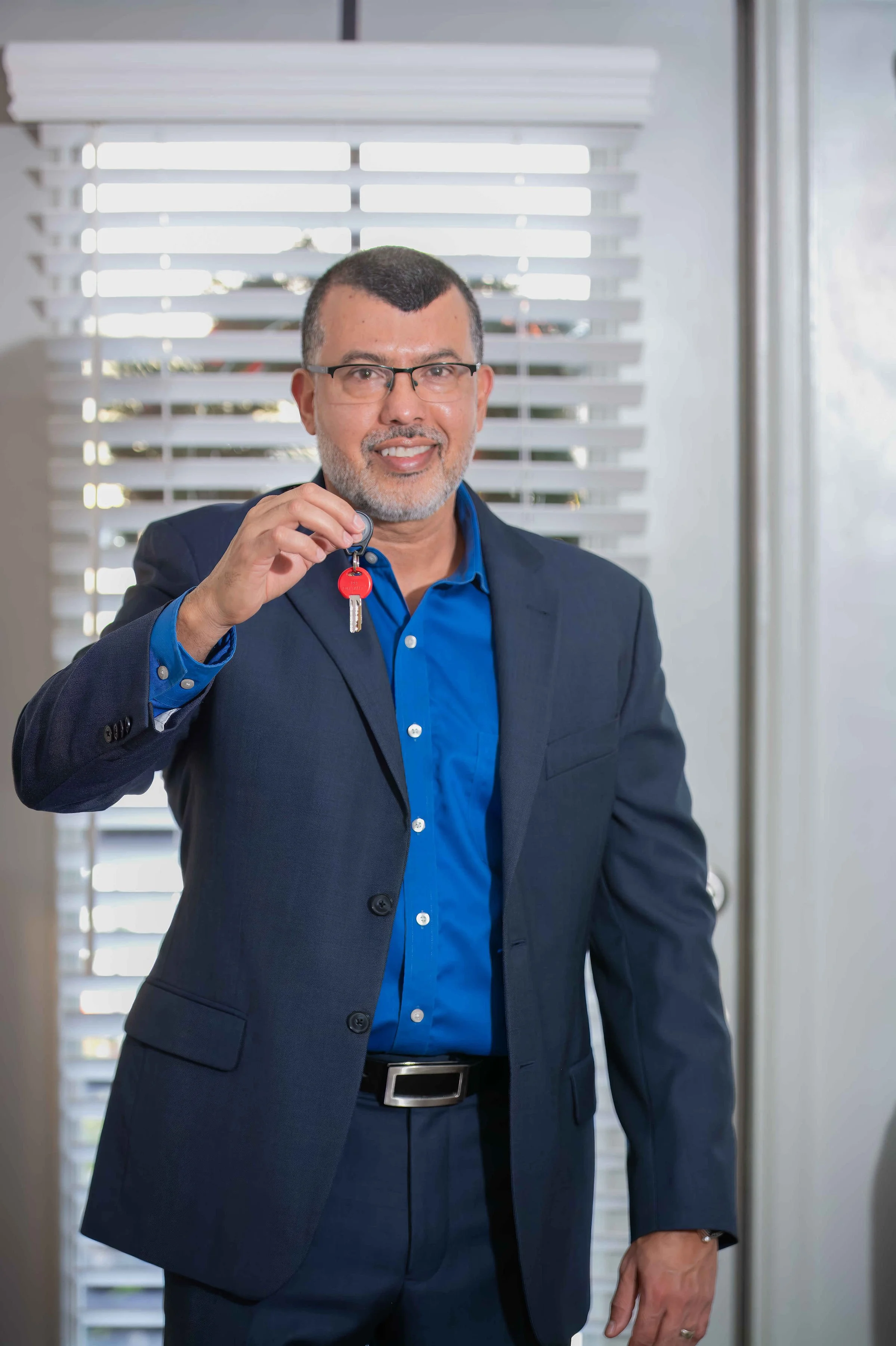 Hombre de negocios con gafas, barba canosa y cabello corto, vestido con traje oscuro y camisa azul, sosteniendo una llave con un llavero rojo, frente a una ventana con persianas blancas.