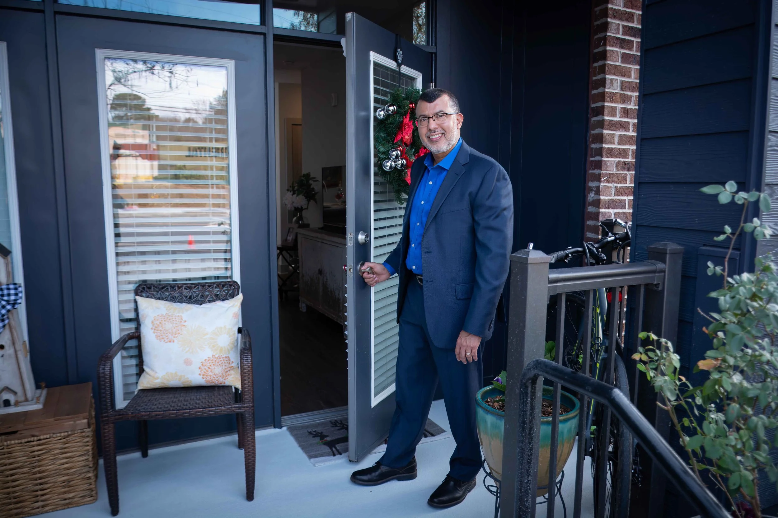 Hombre con traje, gafas y barba, sonriendo en la entrada de una casa decorada para Navidad con una corona y adornos en la puerta, en un balcón con silla, cojín y plantas