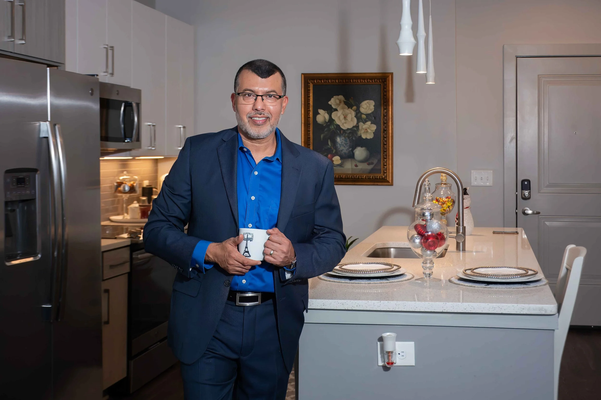 Hombre con gafas y barba, vestido con traje azul, sosteniendo una taza en una cocina moderna con arreglos decorativos y una pintura en la pared.