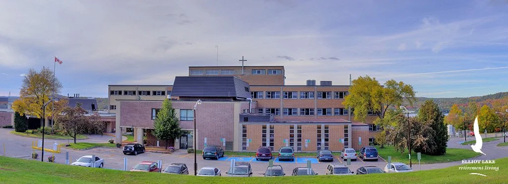 A large multi-story brick retirement living building with ellenic Lake written on the bottom right corner, surrounded by parking lot and trees, under a partly cloudy sky.