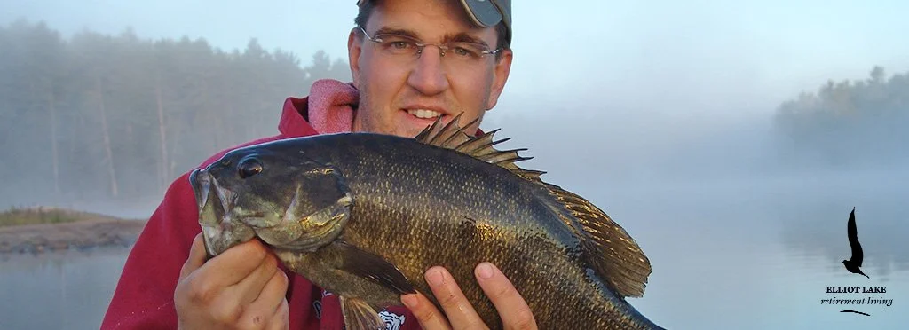 Man in a red jacket holding a large black and green fish near a lakeshore with fog and trees in the background.