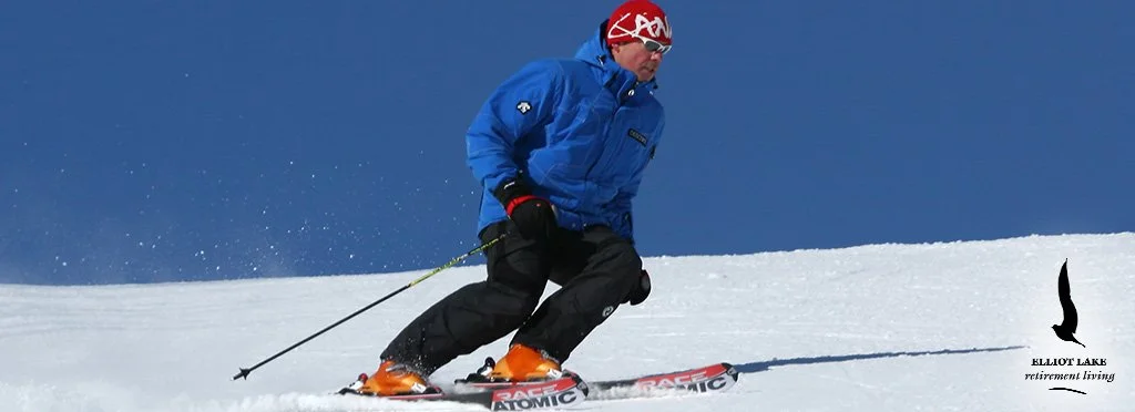 A person skiing down a snowy slope, wearing a blue jacket, black pants, orange ski boots, red helmet with goggles, and holding ski poles. The sky is clear and blue.