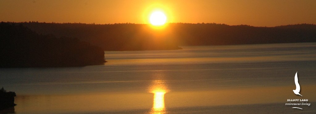 Sunset over a calm lake with a forested shoreline, showing the reflection of the setting sun on the water.