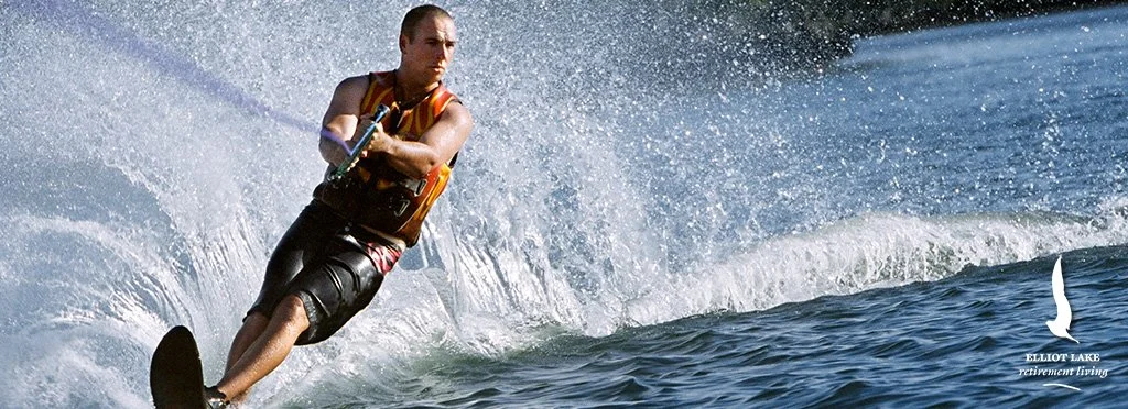 A man waterskiing on a lake with splashing water around him. He is wearing a life vest and shorts, holding onto a tow rope, and leaning to one side as he glides across the water.