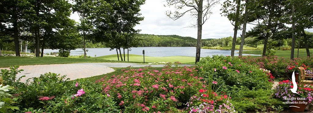 View of a landscaped garden with blooming flowers, a paved patio, and a lake in the background with trees surrounding it.