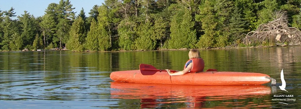 A young girl with blonde hair sitting in an orange kayak on a calm lake surrounded by green trees.