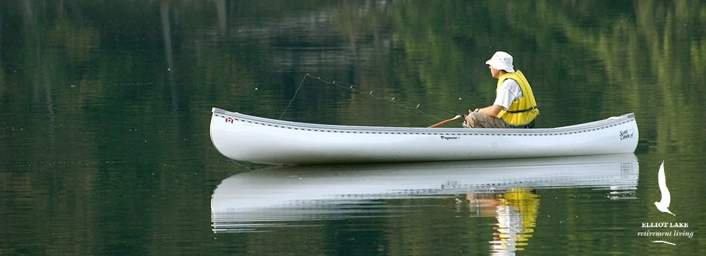 A person in a yellow life jacket and white hat sitting in a white canoe, fishing on a calm lake surrounded by trees.