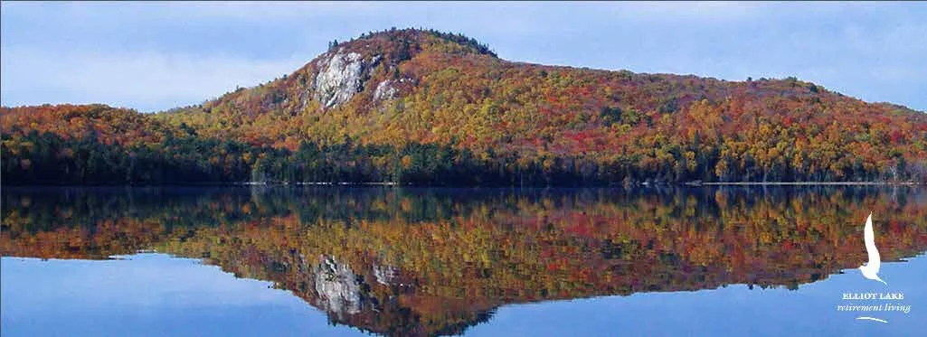 A calm lake reflecting a forested hillside with colorful autumn trees and a rocky peak. There is a small logo in the bottom right corner with a white swan and the text "Kellogg Lake retirement living."