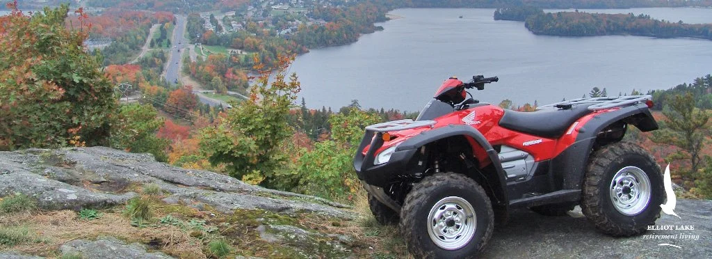 A red ATV parked on a rocky hill overlooking a lake and fall-colored forest.
