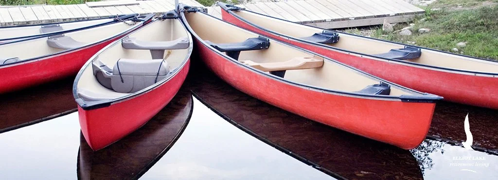 Three red and beige canoes parked side by side on calm water, with a grassy shoreline in the background.