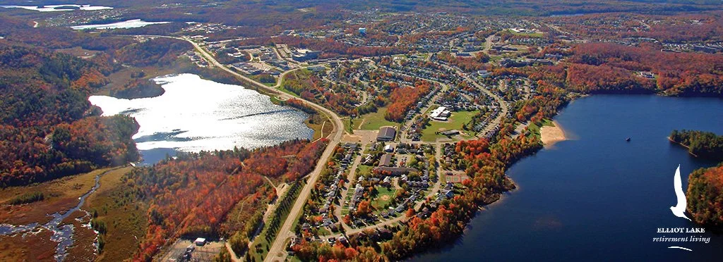 An aerial view of a scenic lakeside area showing a large body of water with surrounding trees in autumn colors, roads, and residential areas.