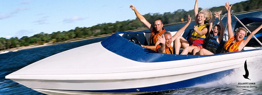Group of five people on a speedboat enjoying a boat ride, wearing life jackets, water splashing behind the boat, trees on shoreline in background.