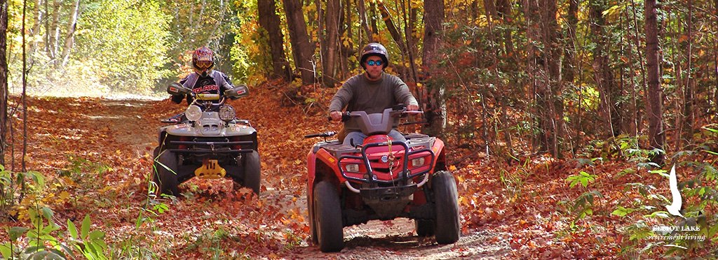 Two people riding all-terrain vehicles on a forest trail surrounded by trees and autumn foliage.