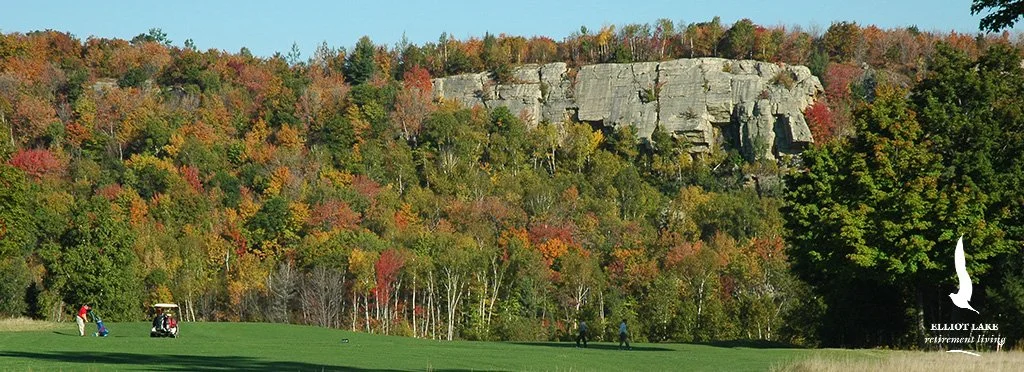 A golf course with four people, one with a golf bag, two walking, and one near a golf cart. The background features a large rocky cliff and trees with autumn foliage.