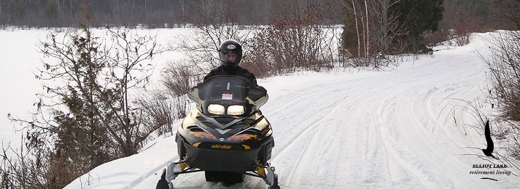 A person wearing a helmet and winter jacket riding a black snowmobile on a snowy trail in a winter landscape.
