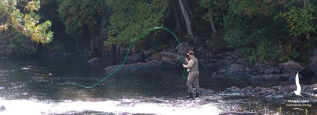 A person fishing in shallow river water, surrounded by trees and rocks.