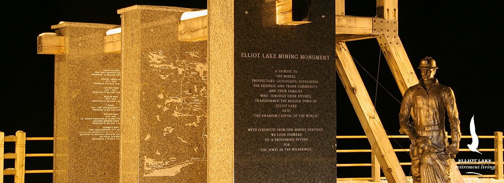 Elliot Lake Mining Monument at night, featuring a bronze miner statue and informational plaques about the history of mining in Elliot Lake, Ontario.