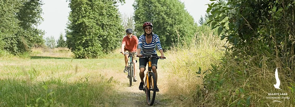Two women cycling on a dirt trail through a green, wooded area during daytime, wearing helmets and casual clothes.