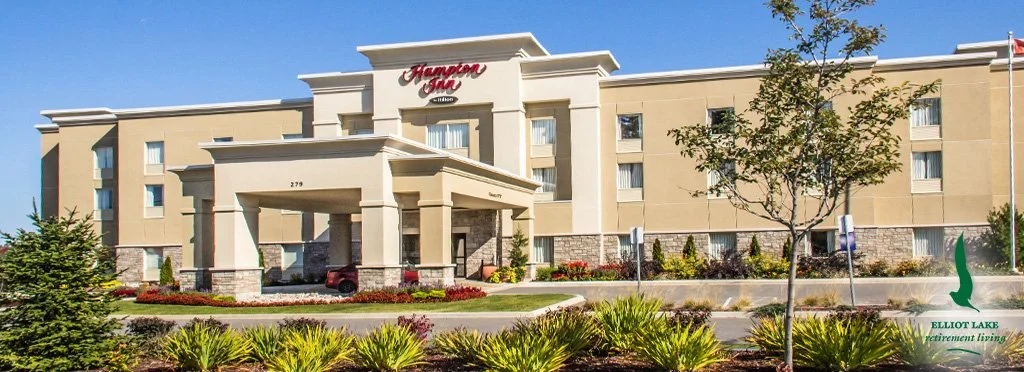 Exterior view of a Hampton Inn hotel with landscaped front yard, some trees, and a blue sky.