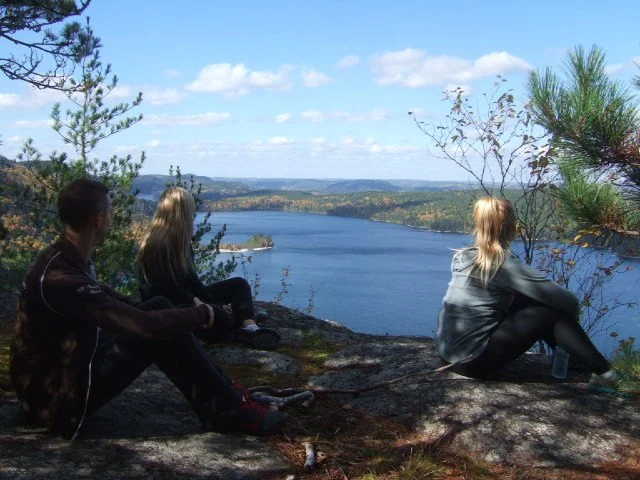 Three people sitting on a rocky ledge overlooking a lake surrounded by trees and hills, with a partly cloudy blue sky.