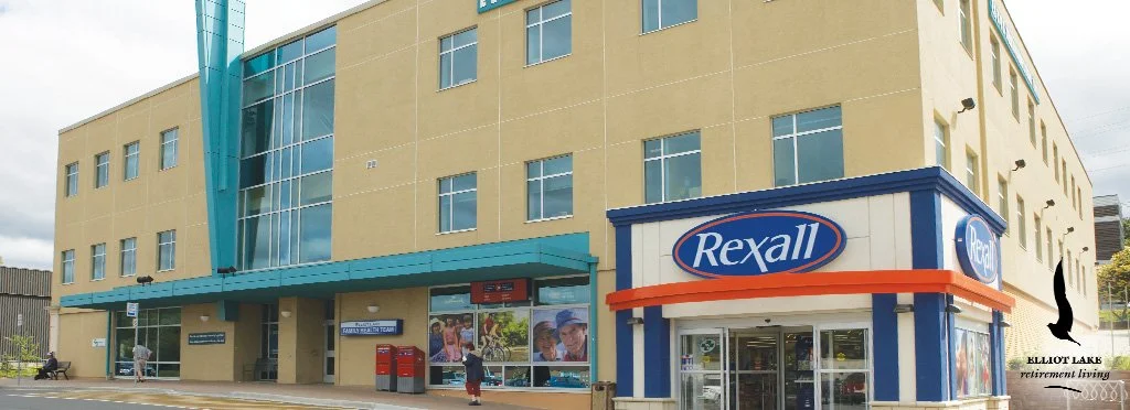 Rexall pharmacy storefront in a multi-story building with a yellow exterior, blue accents, and large windows. People are walking near the entrance, and there are red mailboxes outside.
