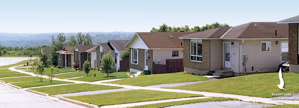 A row of modern suburban houses with manicured lawns and trees along a sidewalk, under a clear blue sky.