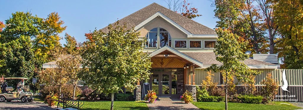 Retirement home with a large, modern house surrounded by autumn trees, a golf cart on the left, and a bench on the lawn, with a sign indicating Elliot Lake Retirement Living.