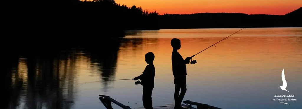 Two children fishing at sunset on a calm lake with orange sky, silhouetted against the water and trees.