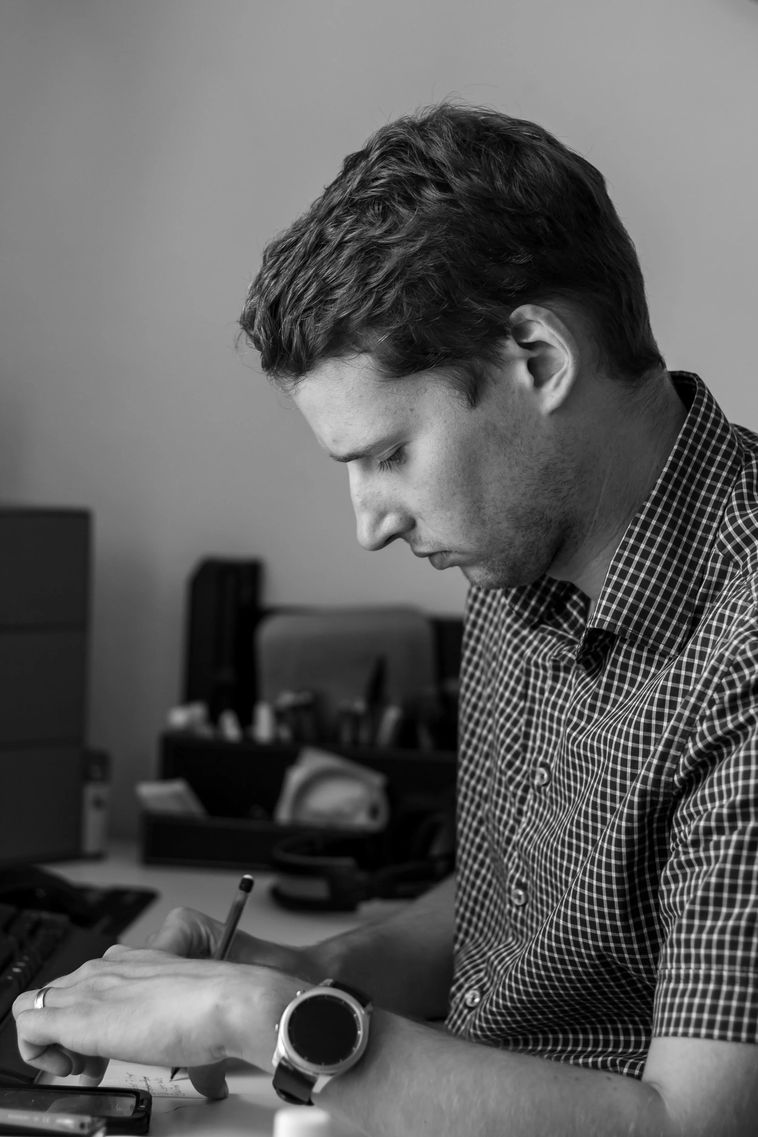 A man with dark, wavy hair wearing a checkered shirt and smartwatch, sitting at a desk, writing with a pen, in an office space with computer monitors and office supplies in the background.