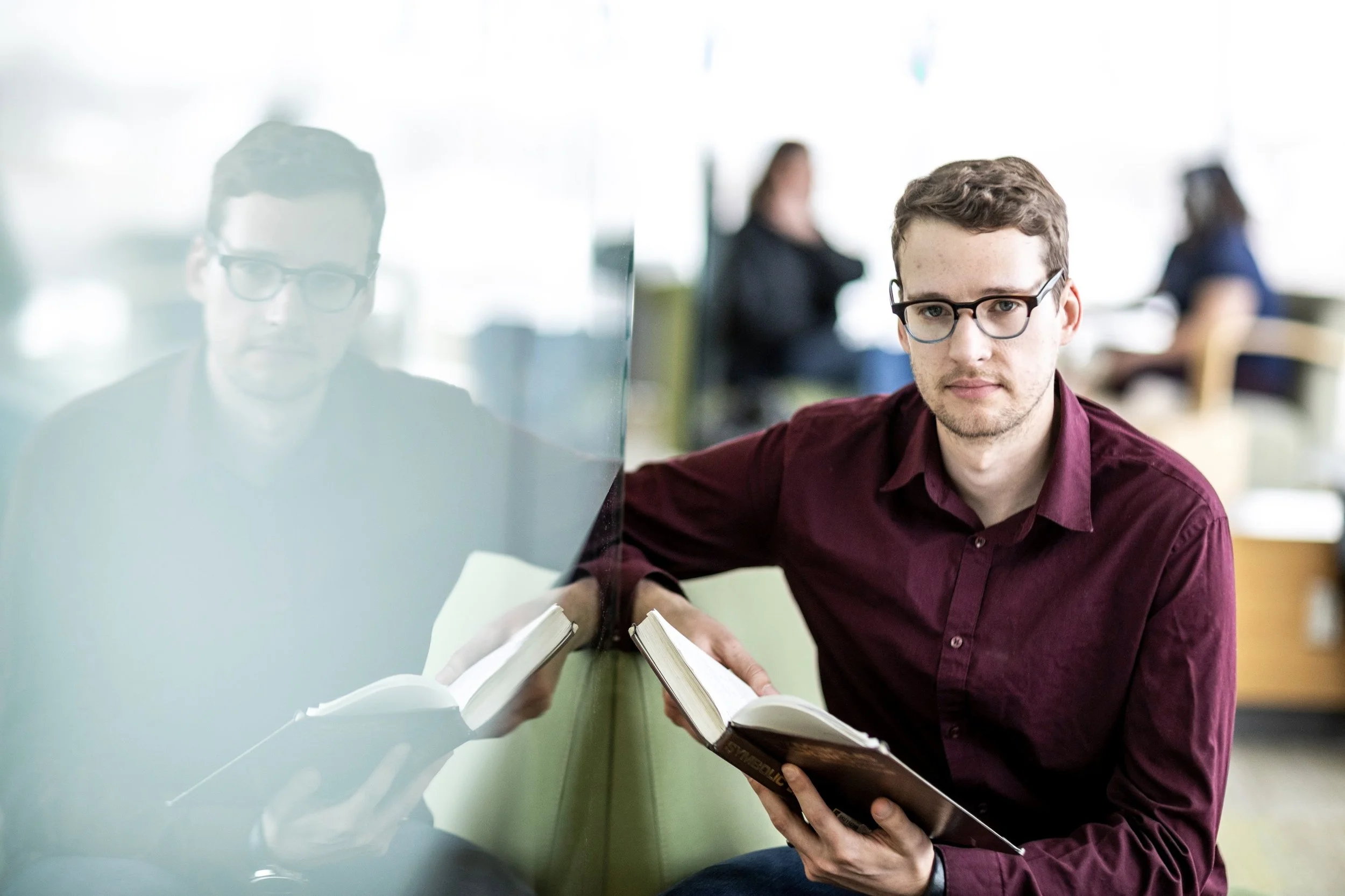 Young man with glasses studying a book in a modern, well-lit indoor space, with colleagues in the background.