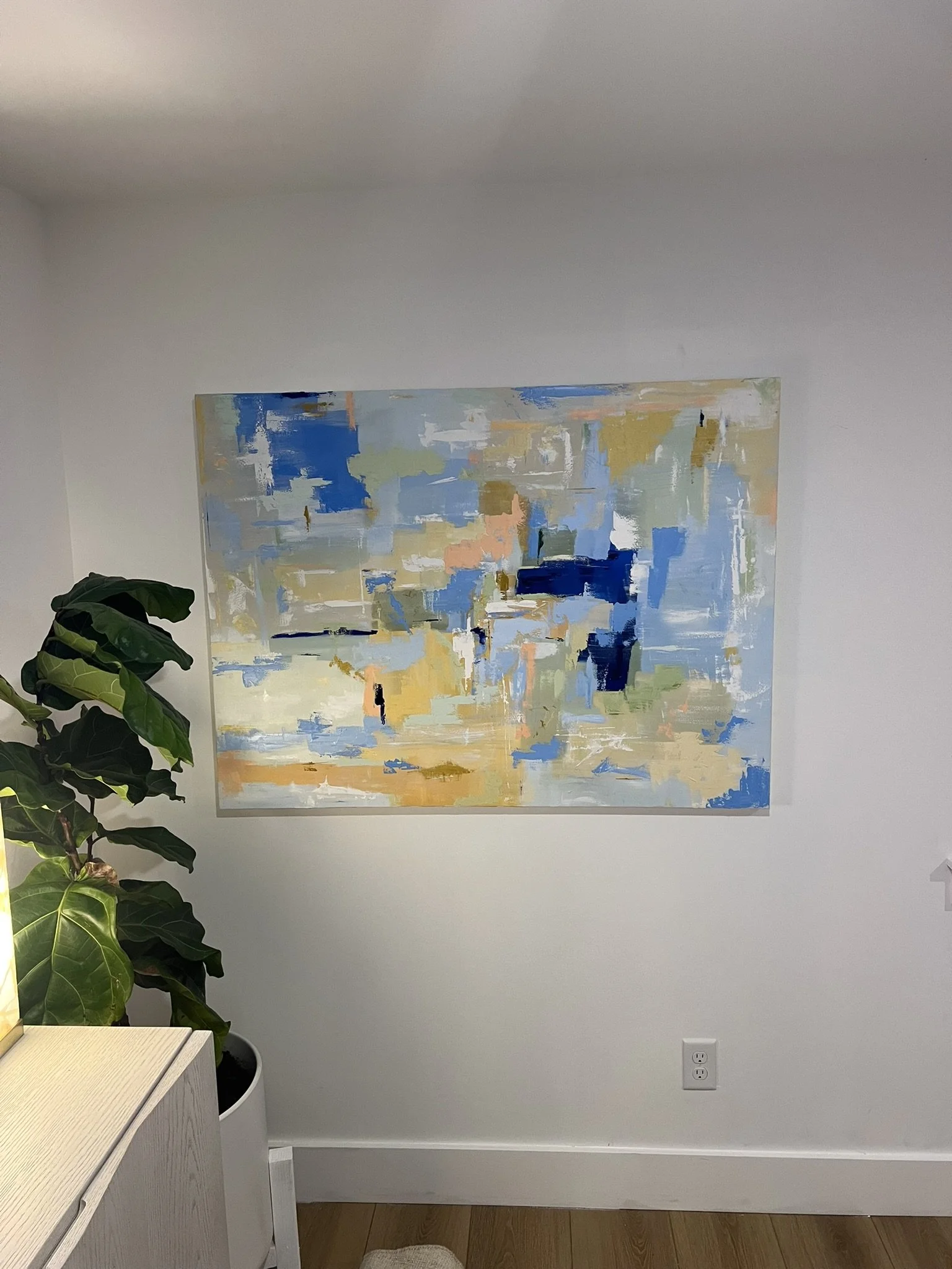 Interior view of a room with a white wall and a large abstract painting with blue, yellow, beige, and white brushstrokes. A green plant with large leaves is on the left side, and part of a light-colored wooden cabinet with a drawer is visible in the bottom left corner. There is a double electrical outlet on the wall near the floor.