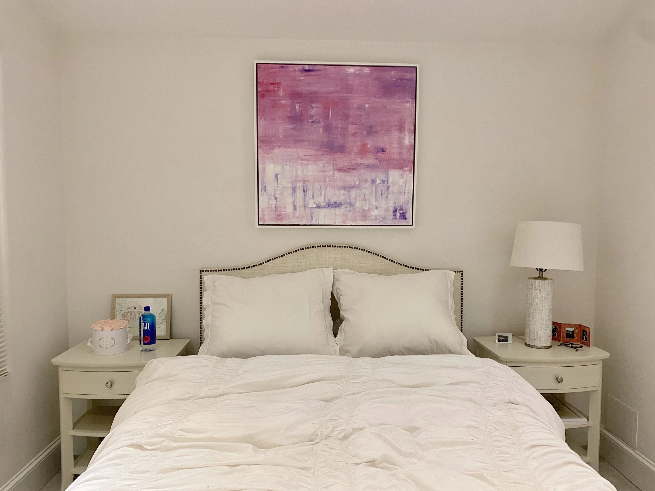 A bedroom with a white bed, bedside tables, and lamps, framed artwork above the bed, and various decorative items on the tables.