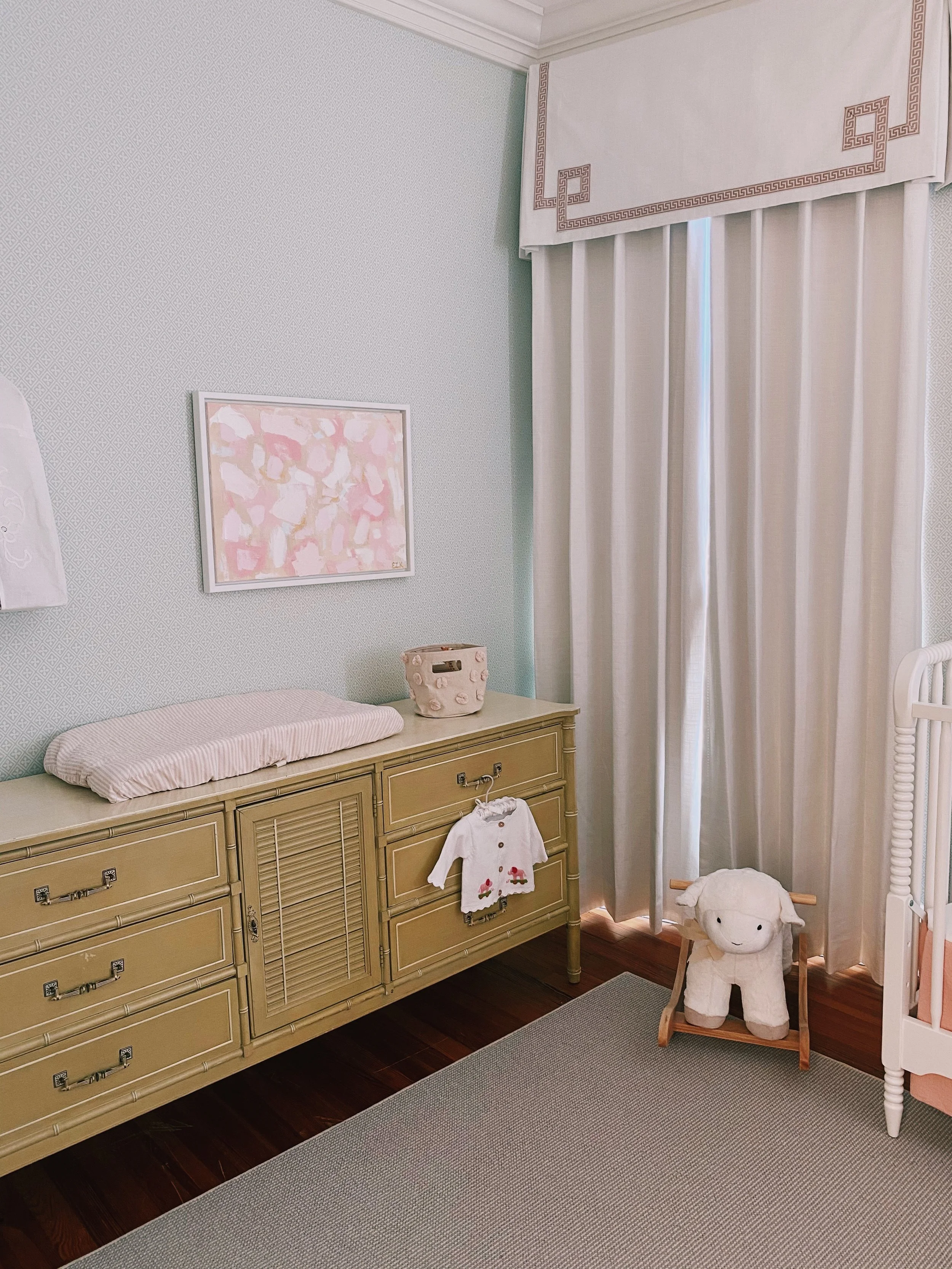 A nursery corner with a beige dresser, a pink and white changing pad, framed pink abstract art, a white plush sheep, a small dollhouse, and cream curtains in a room with wooden flooring.