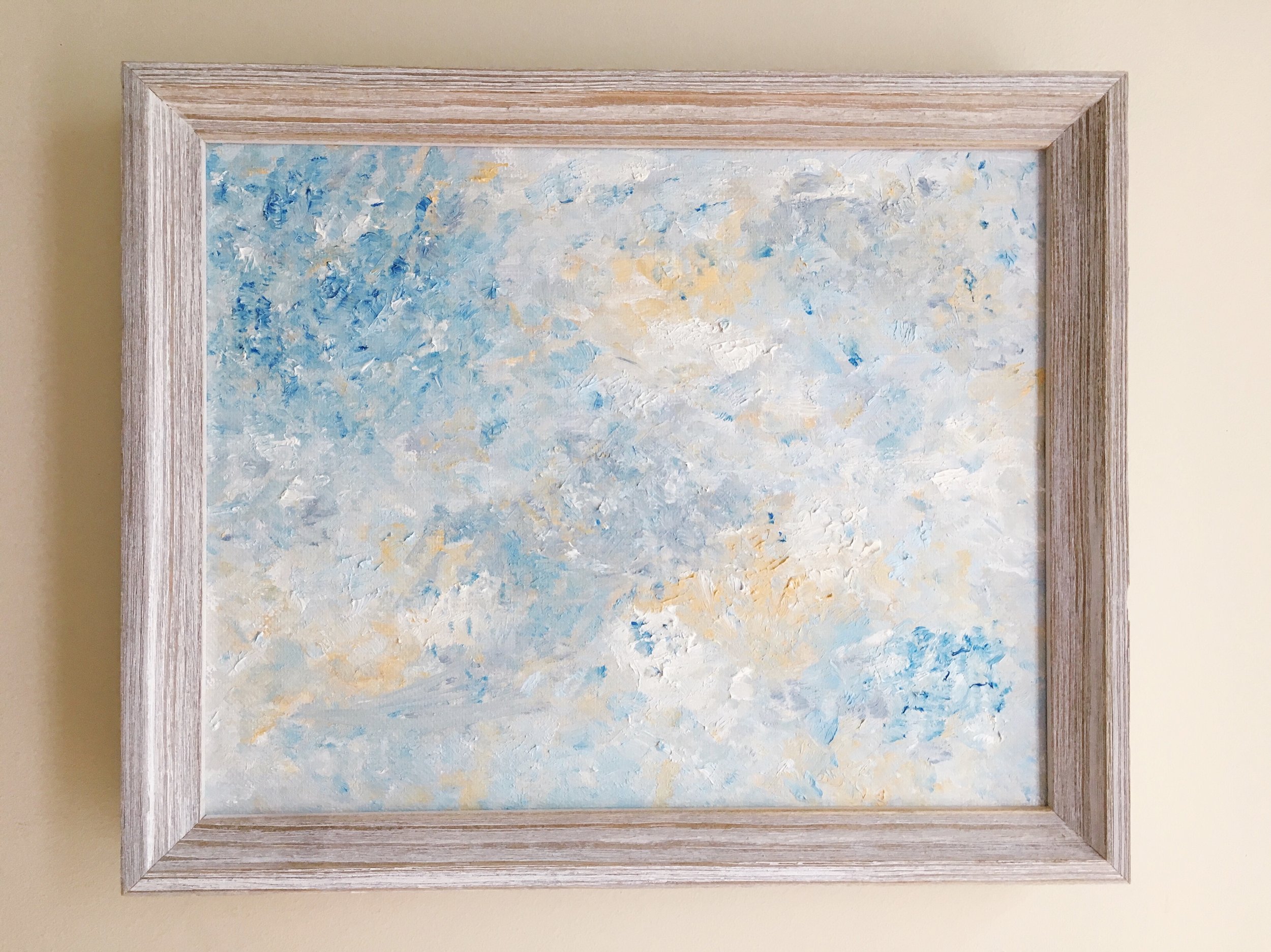 An abstract painting with shades of blue, white, and yellow, framed by a light wood frame, hanging on a light-colored wall.