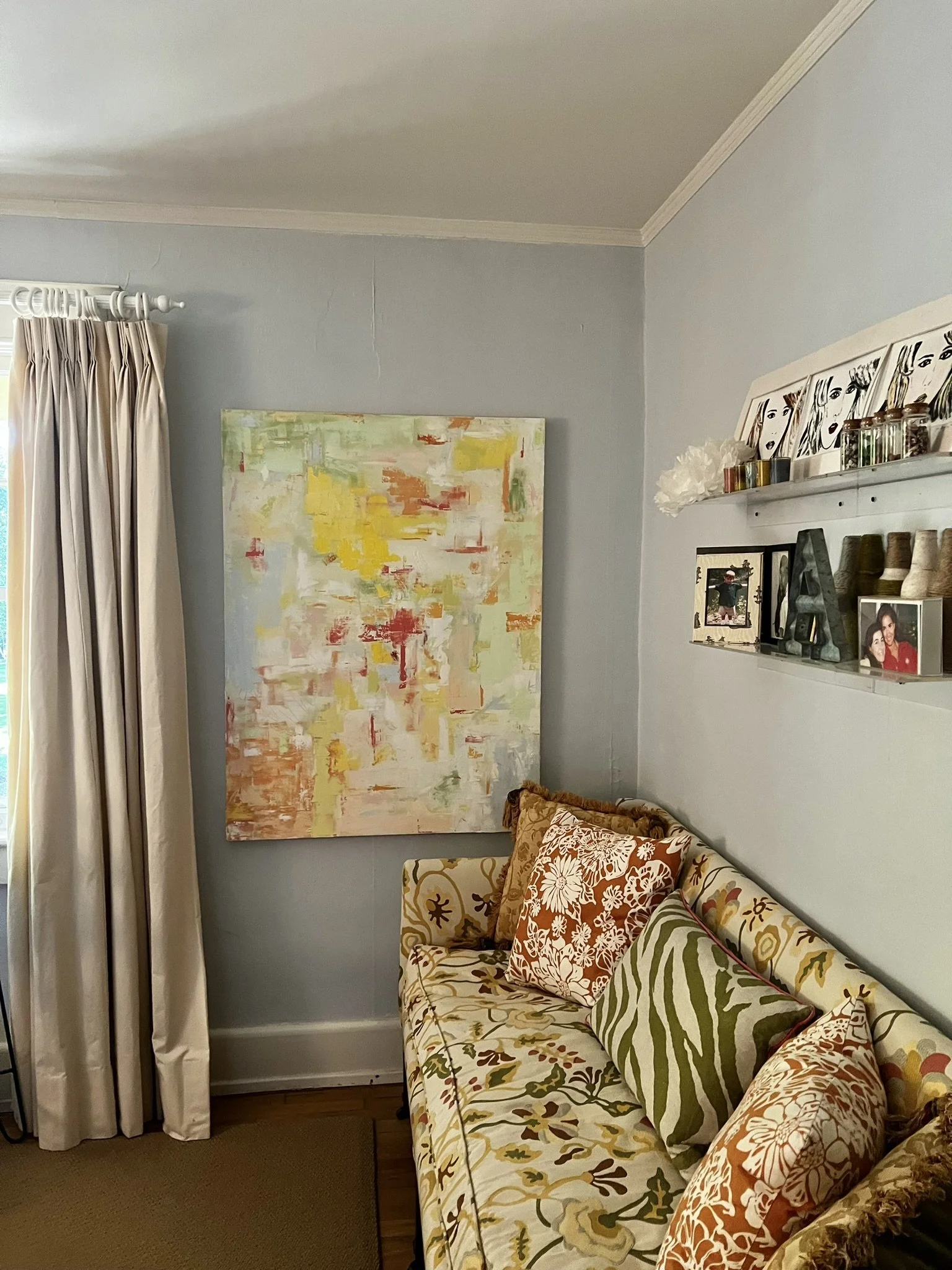 A living room corner with a patterned sofa, decorative pillows, a colorful abstract painting on a light blue wall, and a white shelf with framed pictures, candles, and jars.