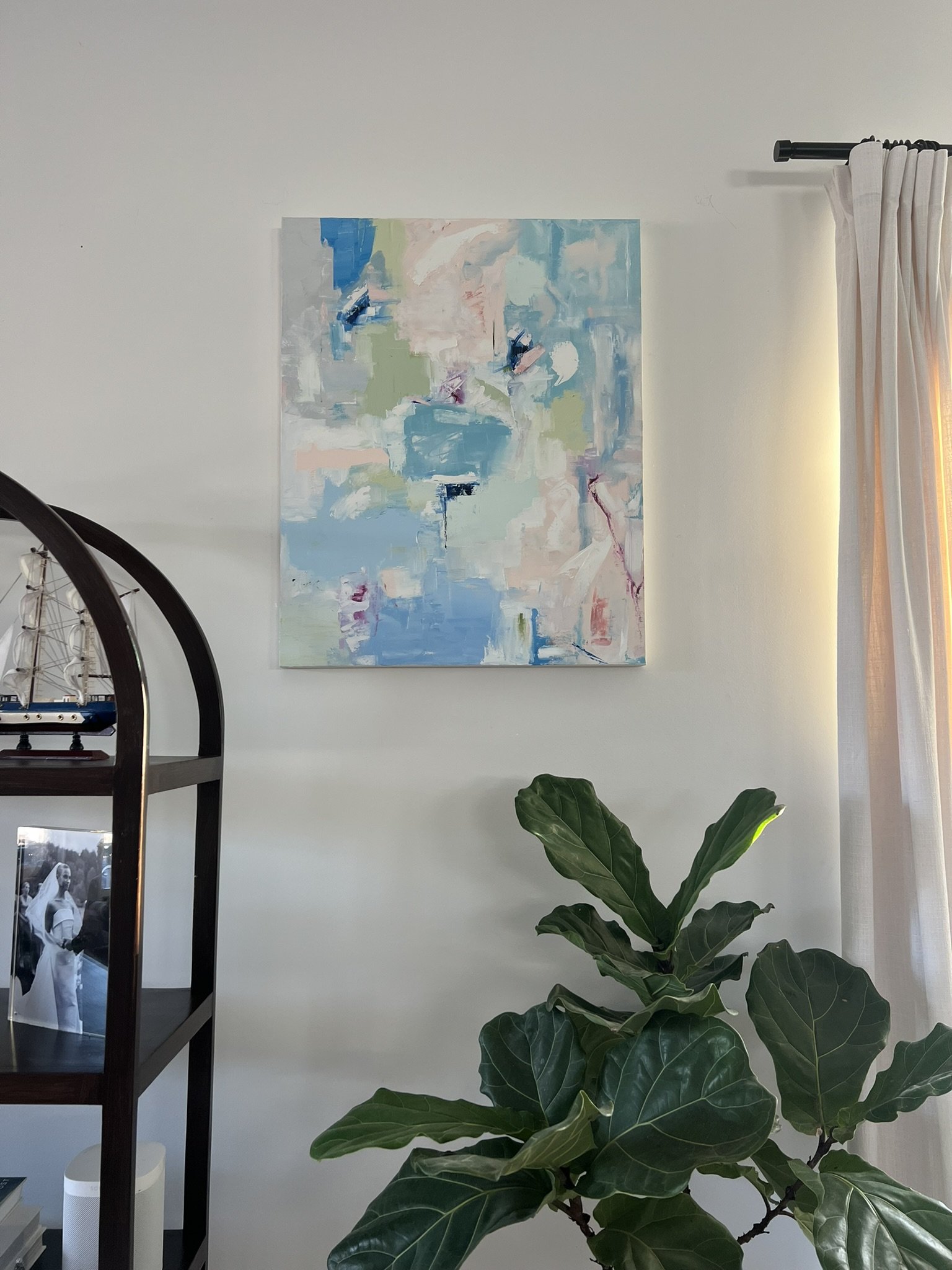 Interior of a room with a white wall, a framed abstract painting with blue, green, white, and pink colors, a large green leafy plant, a black bookshelf with framed black-and-white photographs, and a white smart speaker on a lower shelf. There are also curtains near a window on the right side.