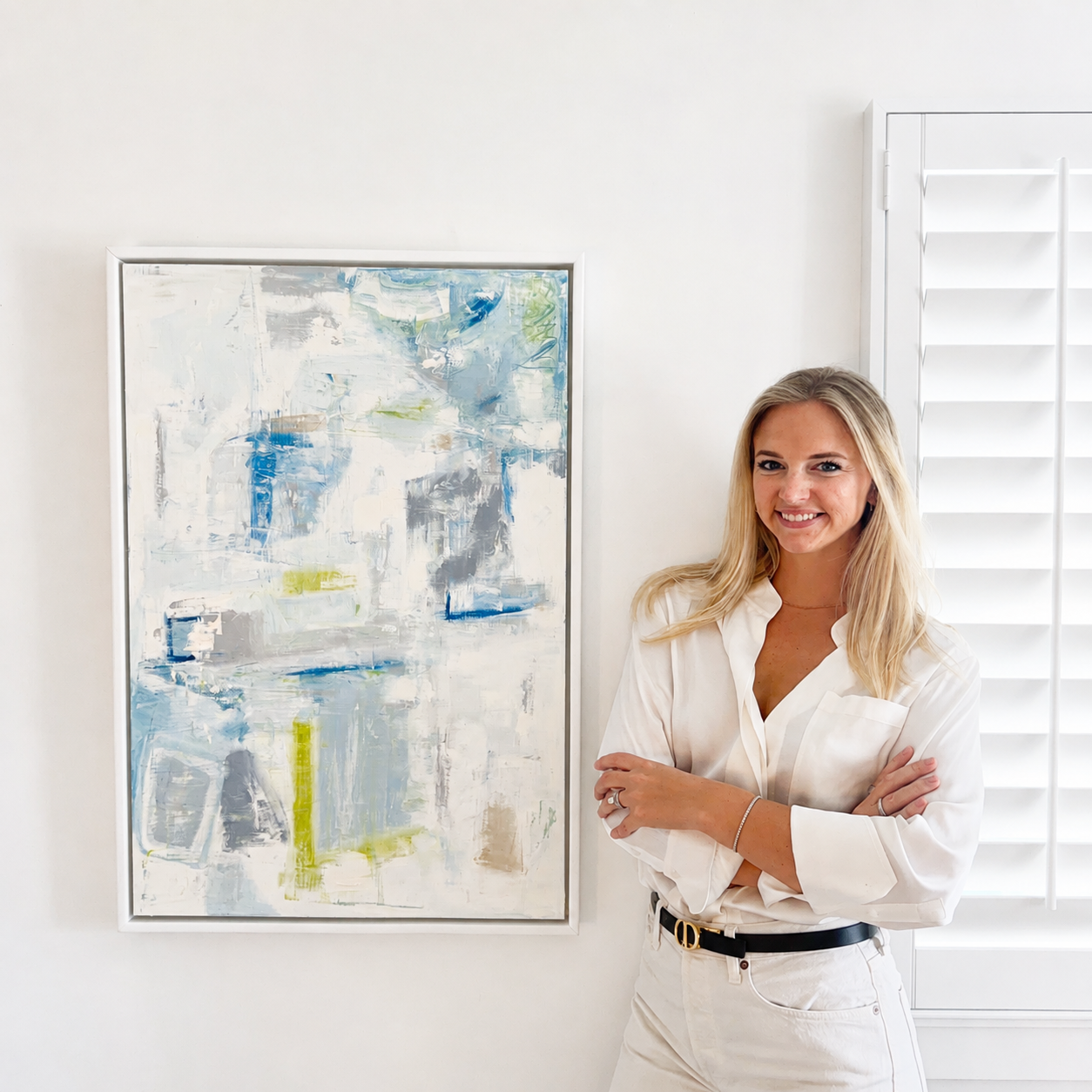 A woman with long blonde hair smiling and crossing her arms, standing in front of an abstract painting on a white wall.