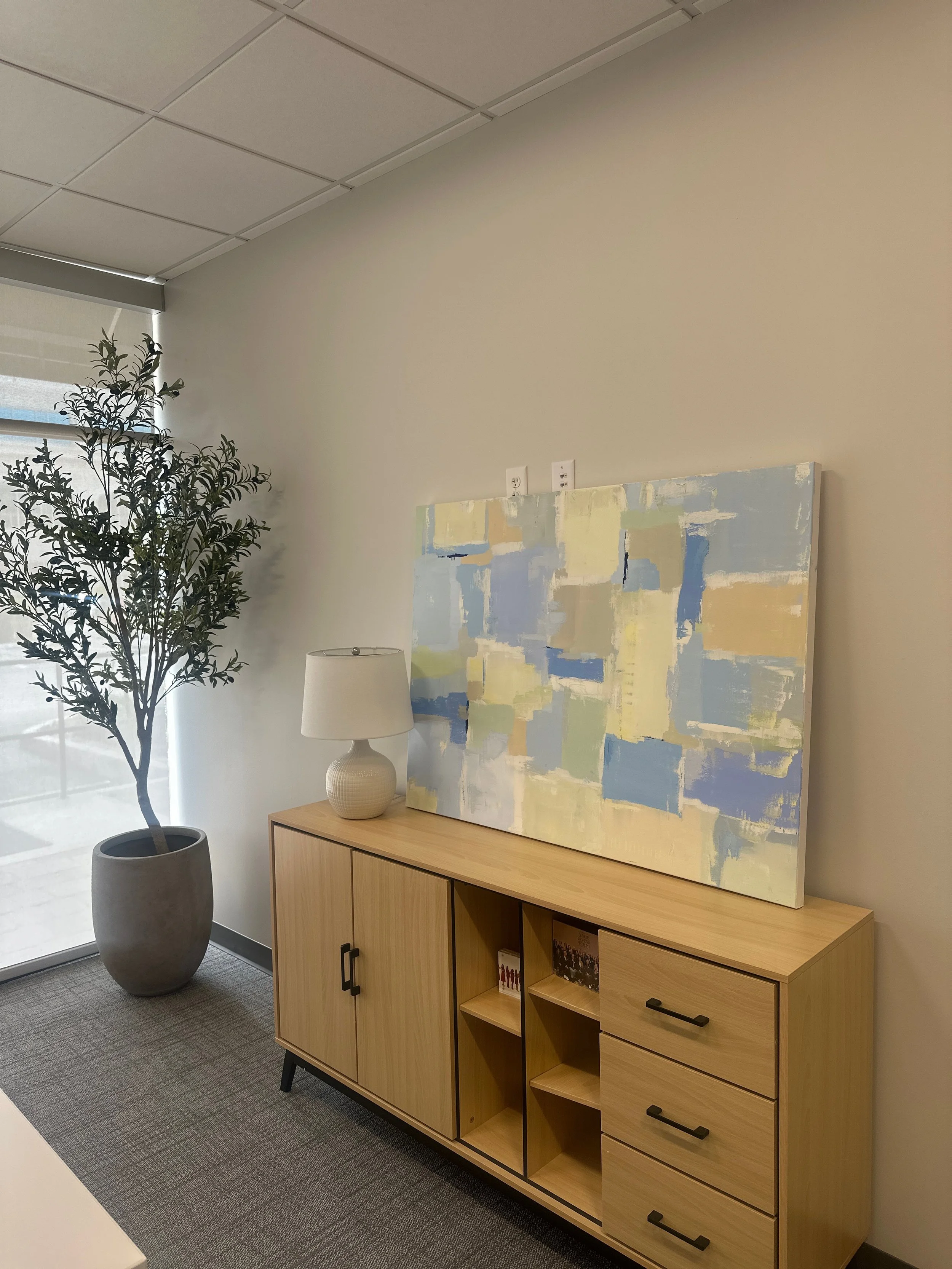 Interior office room with a tall potted plant near the window, a wooden sideboard, a white table lamp, and an abstract painting on the wall.