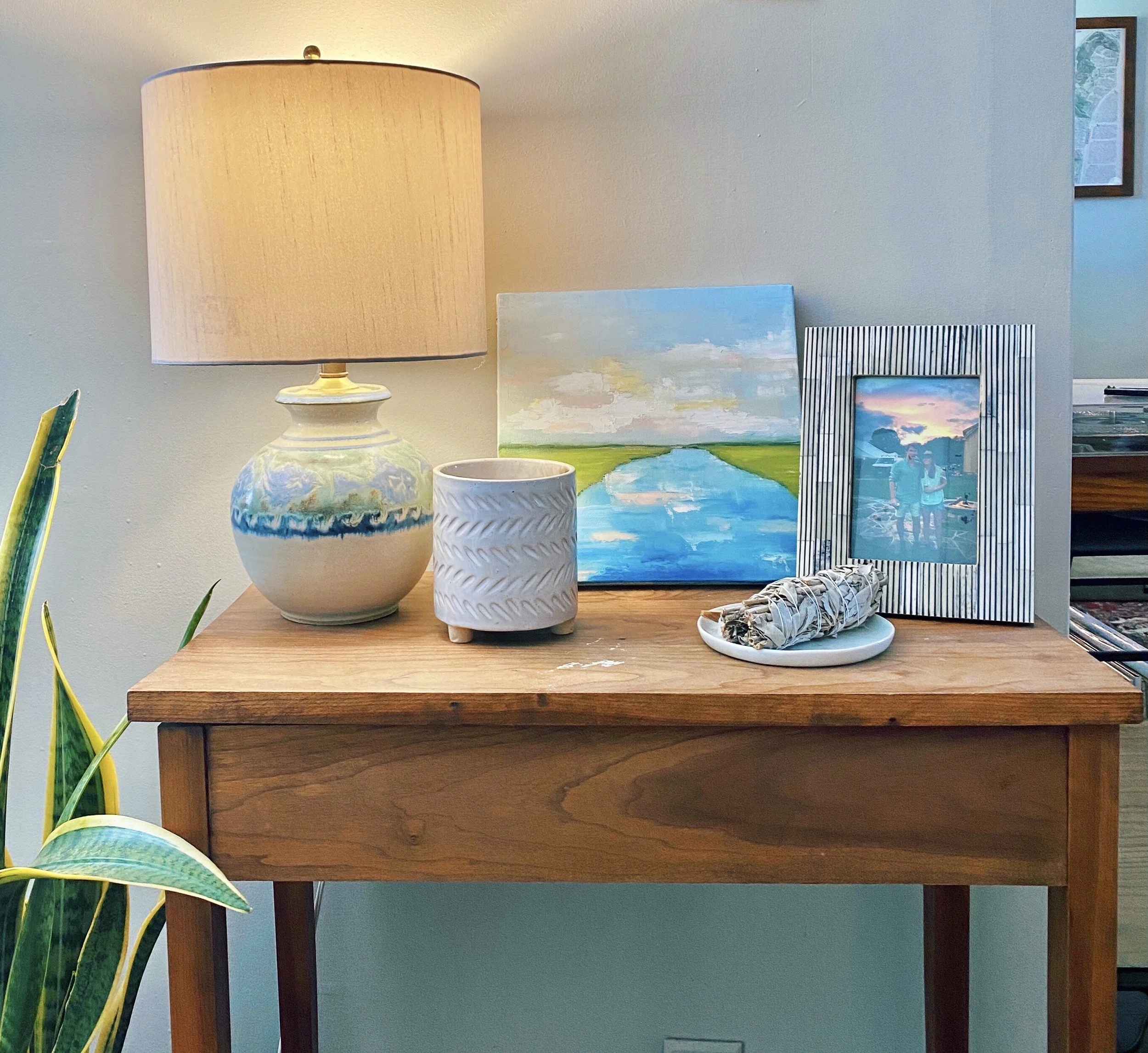 A wooden table with a beige lampshade, two ceramic vases, a rolled-up newspaper, a framed mirror reflecting a sunset, and paintings of outdoor landscapes against a light-colored wall.