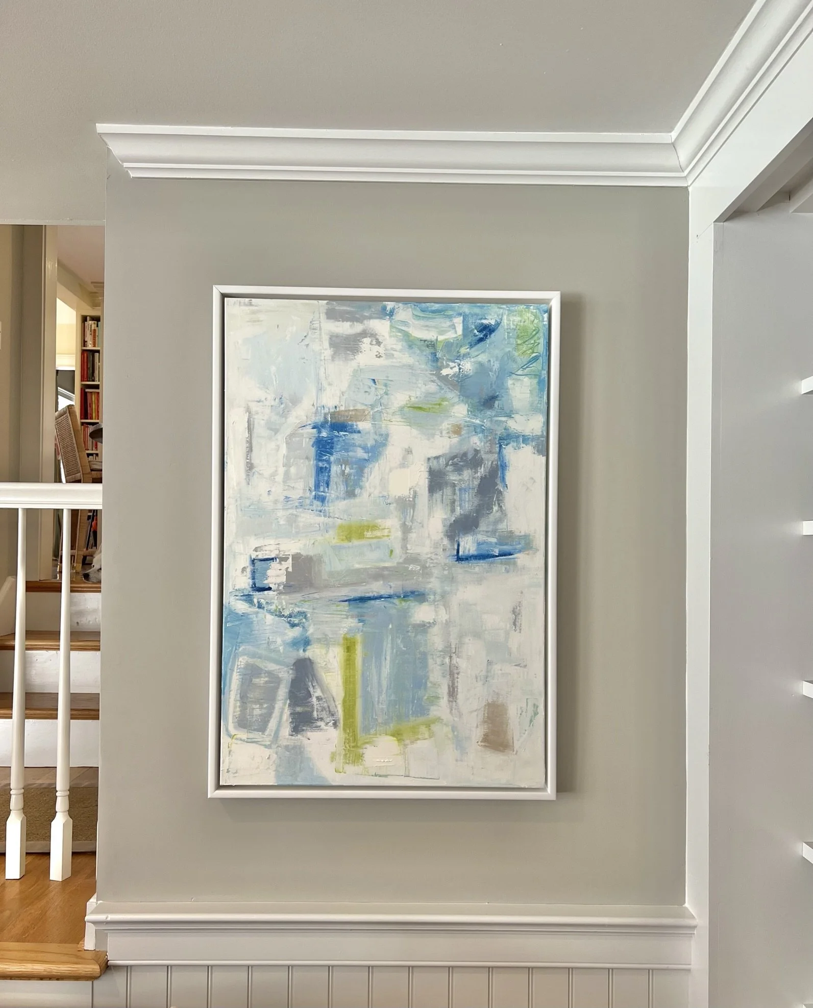 An abstract painting with blue, green, gray, and white brushstrokes in a white frame hanging on a beige wall.