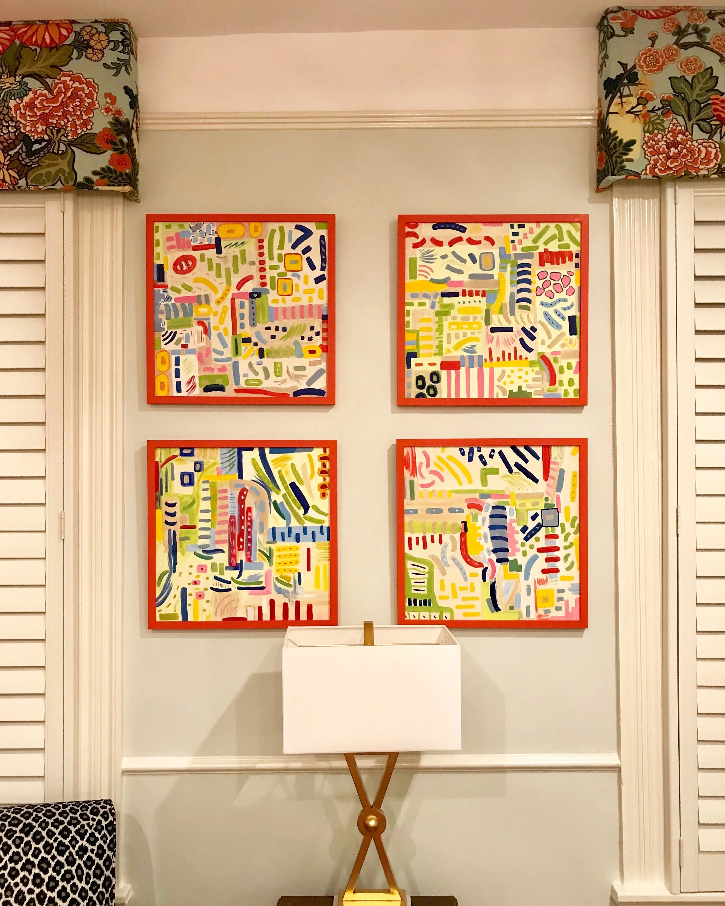 Interior of a room with colorful abstract paintings framed in red hanging on a white wall between two decorative window shutters. A small wooden table with a lamp is positioned below the paintings.