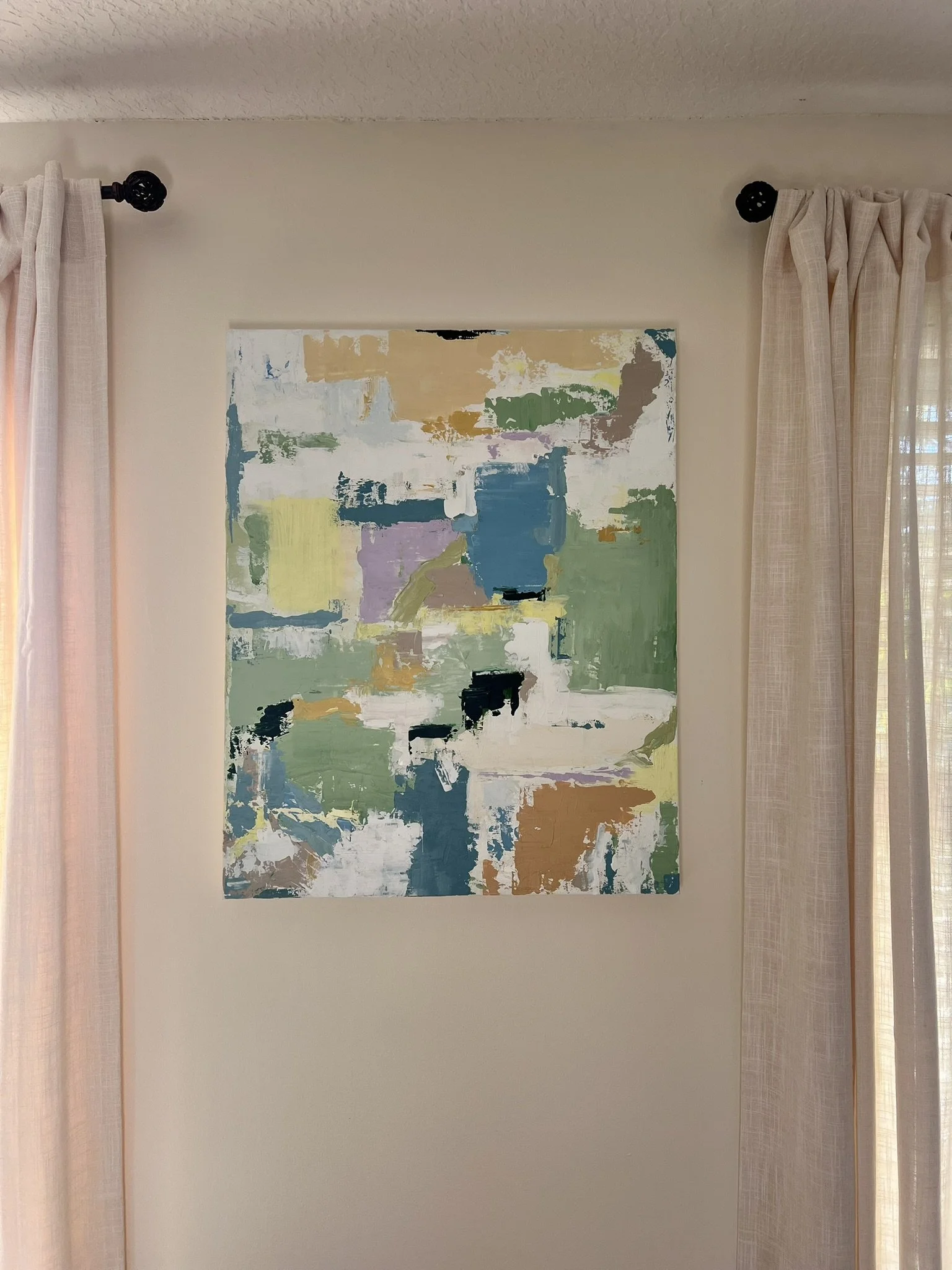 An abstract painting with various colors including blue, green, yellow, purple, white, and beige hanging on a beige wall between pink curtains.