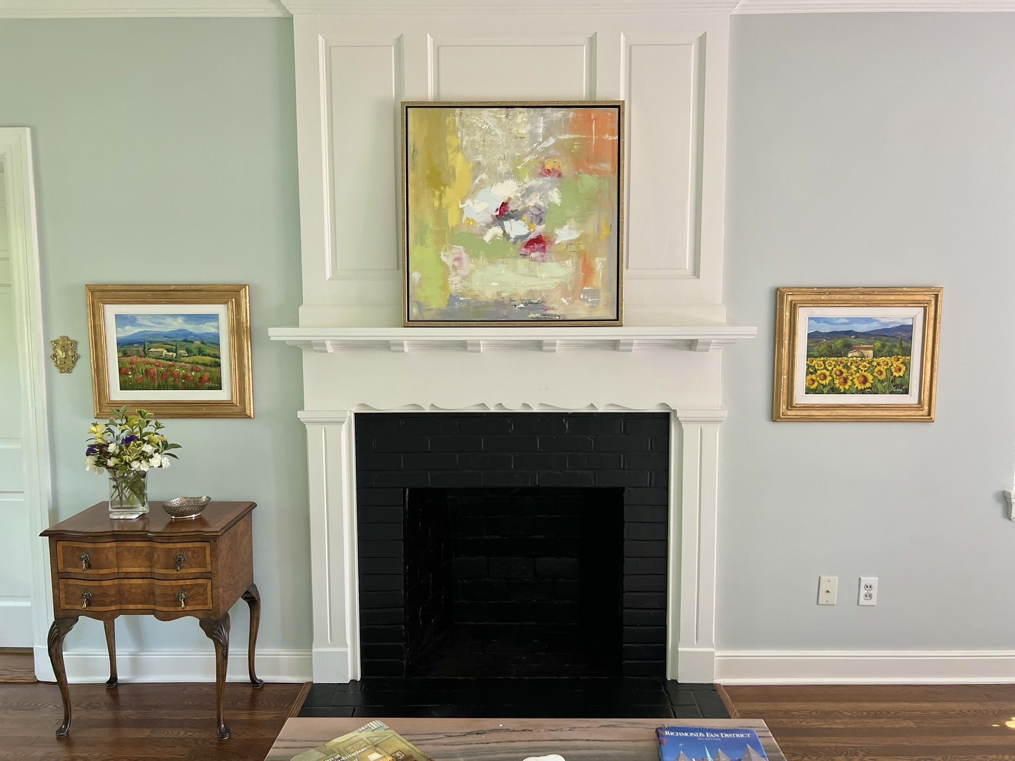 A decorated living room with a white fireplace, two landscape paintings in gold frames on either side, a wooden side table with a colorful flower arrangement, and a colorful abstract painting above the fireplace.
